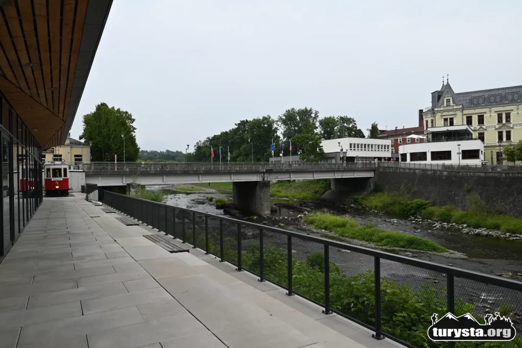 Widok z dystansu na czerwony tramwaj stojący na deptaku w Cieszynie, z malowniczą rzeką Olzą w tle.