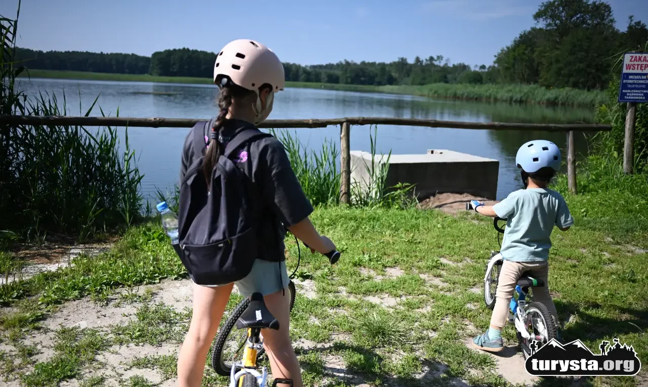 Dzieci stojące na pomoście nad stawem, patrzące na wodę; oboje z rowerami.