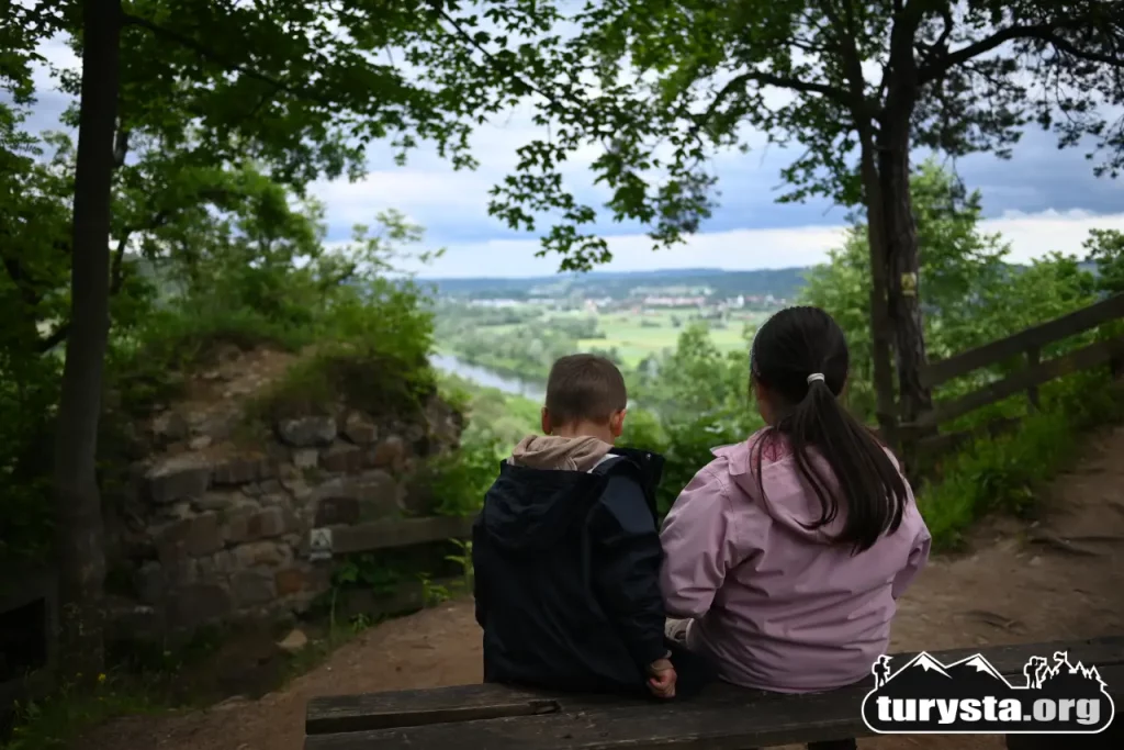 Dwoje dzieci siedzących na drewnianej ławce na wzgórzu zamkowym w Melsztynie, podziwiających zieloną panoramę Doliny Dunajca.