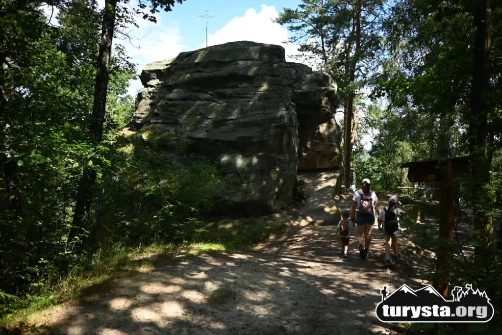Turyści idący leśną ścieżką w kierunku monumentalnej Skałki z Krzyżem w Rezerwacie Skamieniałe Miasto w Ciężkowicach.