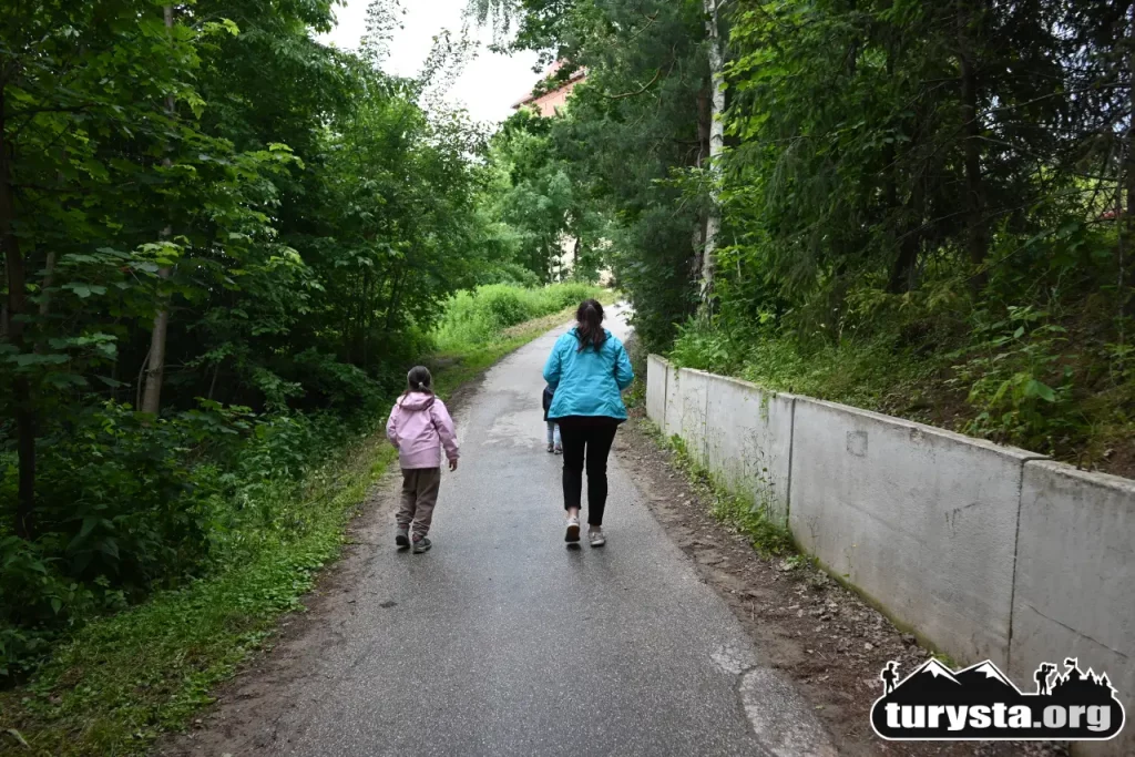 Rodzina spacerująca gładką, asfaltową ścieżką pod górę w stronę zamku w Melsztynie, otoczona zielonymi drzewami.