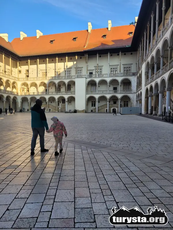 Rodzic z małym dzieckiem spacerujący po brukowanym dziedzińcu arkadowym na Zamku Królewskim na Wawelu. Piękna renesansowa architektura.
